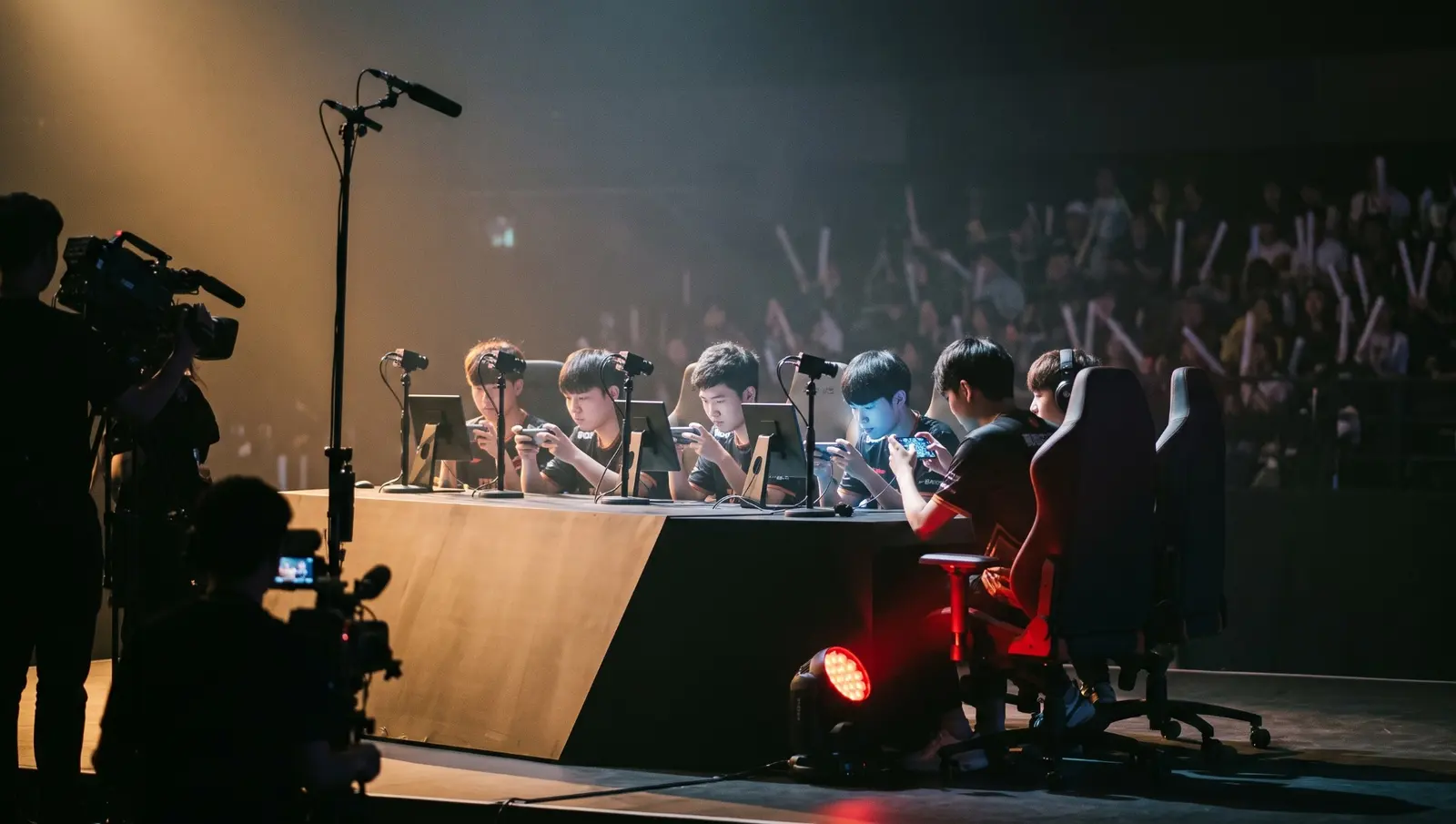 Players at a mobile esports tournament desk, phones in competition stands, a camera crew in silhouette and a lit arena crowd behind them